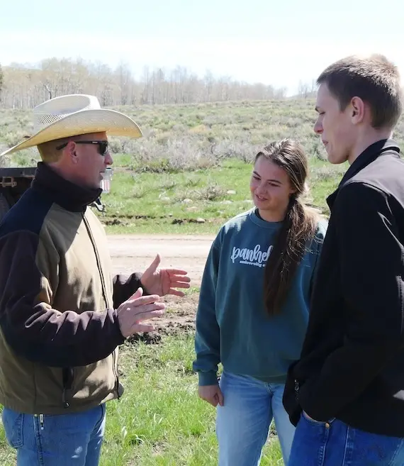 middle-aged man wearing cowboy hat gestures with his hands to explain a concept to two young people in a rangeland setting