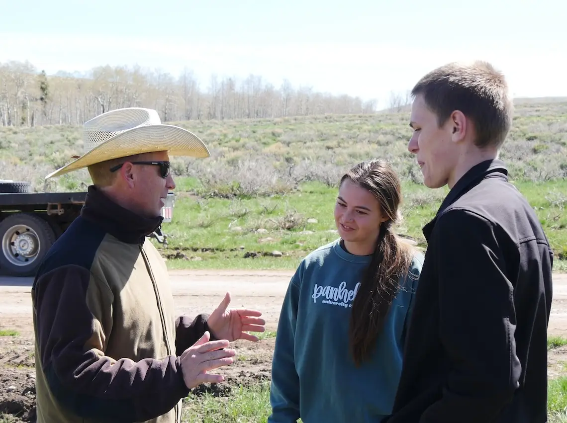 middle-aged man wearing cowboy hat gestures with his hands to explain a concept to two young people in a rangeland setting