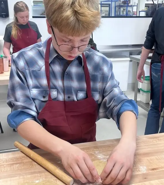boy wearing apron and glasses forms a homemade tortilla on a wooden-topped counter