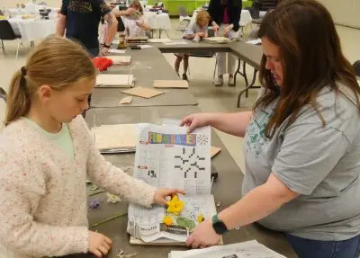 4-H educator holds open a sheet of newspaper on top of a wooden flower press while child sets flower blossoms and leaves inside