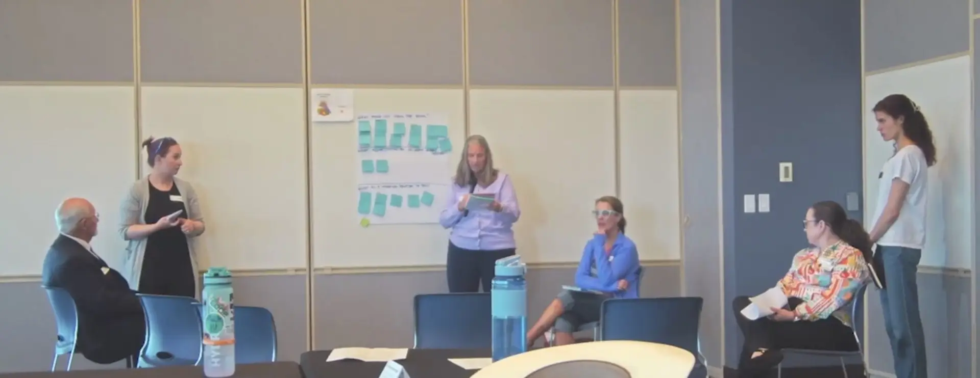 two people stand at the front of a room in front of a whiteboard with sticky notes affixed to it while several seated participants listen to the speakers