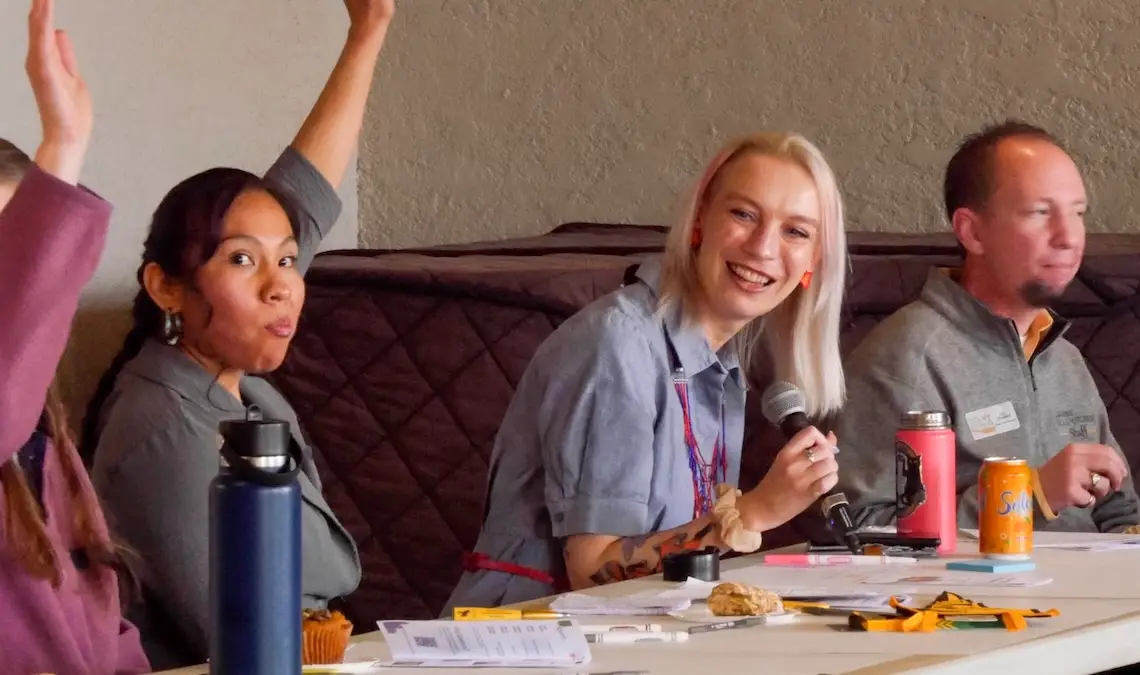 several people seated at a table participate in a panel discussion. One person is holding the mike and two are raising their hands.