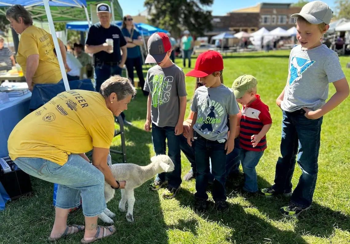 woman holding lamb bends down to show a lamb to four children in a grassy area surrounded by small vendor tents