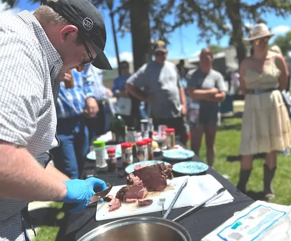man wearing black UW hat and blue food safety gloves cuts a sample of cooked lamb at an outdoor serving table