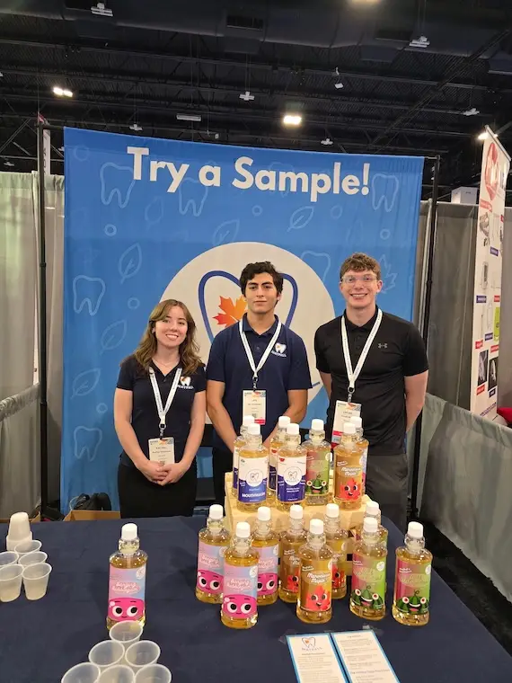Three young adults stand at a booth with samples of Maypall mouthwash products.