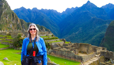 A woman in a blue jacket stands in front of the stone ruins and steep green mountains of Machu Picchu, Peru.