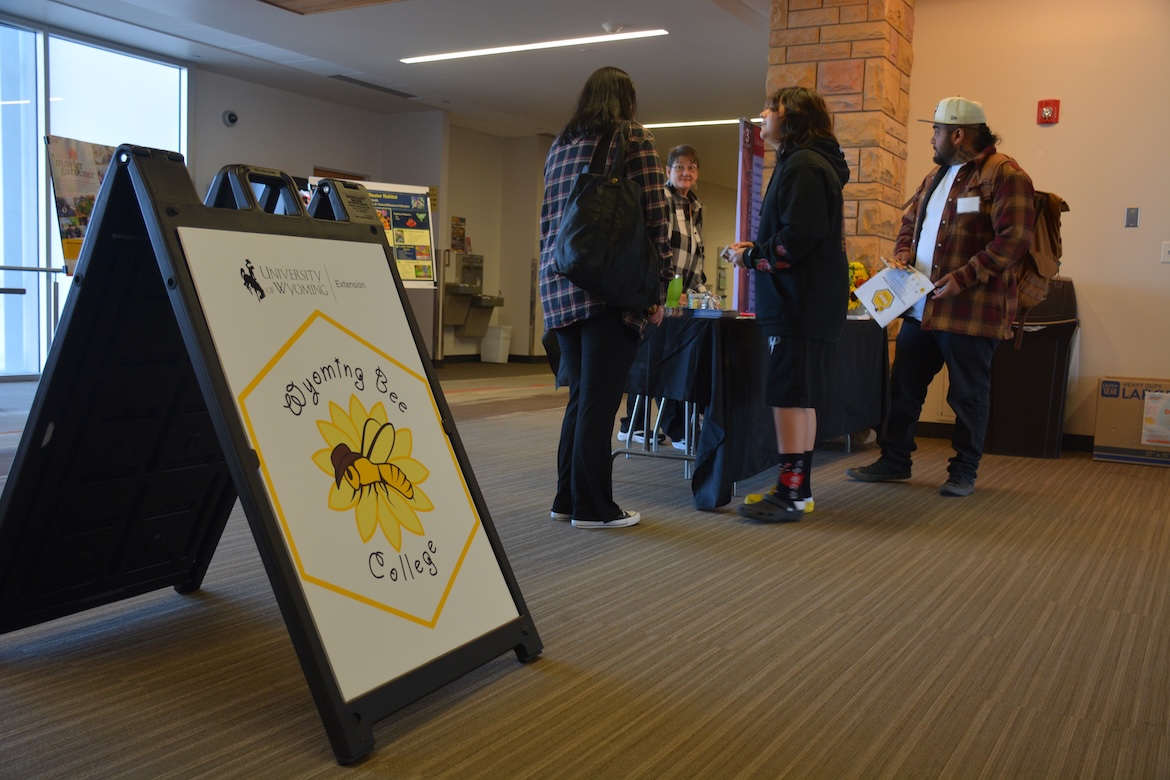 A poster reading "Wyoming Bee College" in the foreground while several people check in at a booth.