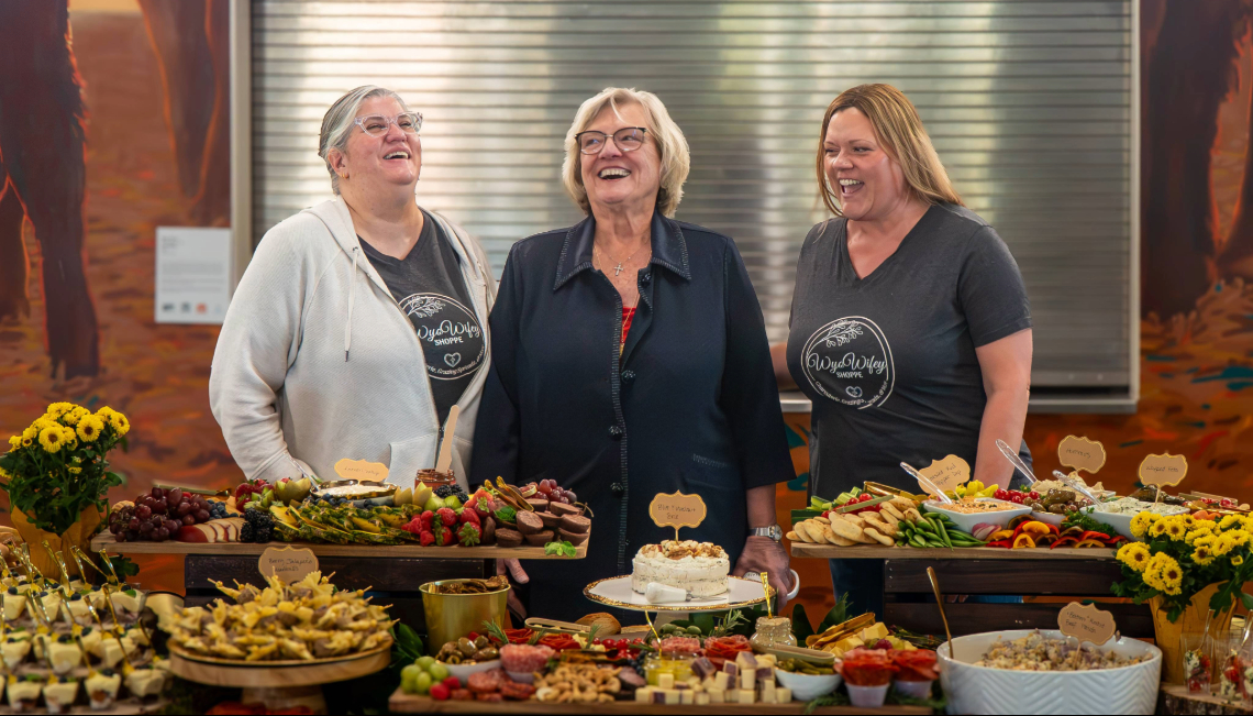 Mary Martin laughs with two women behind a table filled with food and flowers at her retirement celebration.