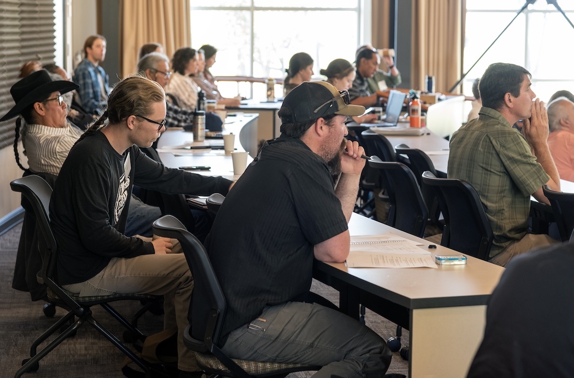 people seated at tables face forward or lean down to take notes during a presentation in a windowed meeting room