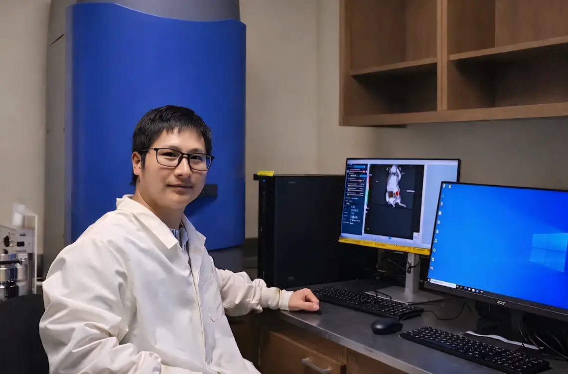 Kiet Tran, wearing labcoat, sits in front of two monitors, one showing a diagram of a mouse