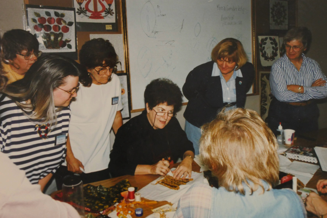 A group of women gathers around a table as an instructor demonstrates a quilting technique.