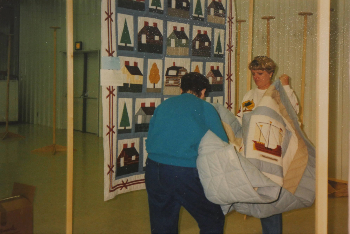 Two women hang a large quilt at an indoor quilting event, with another quilt displayed in the background.