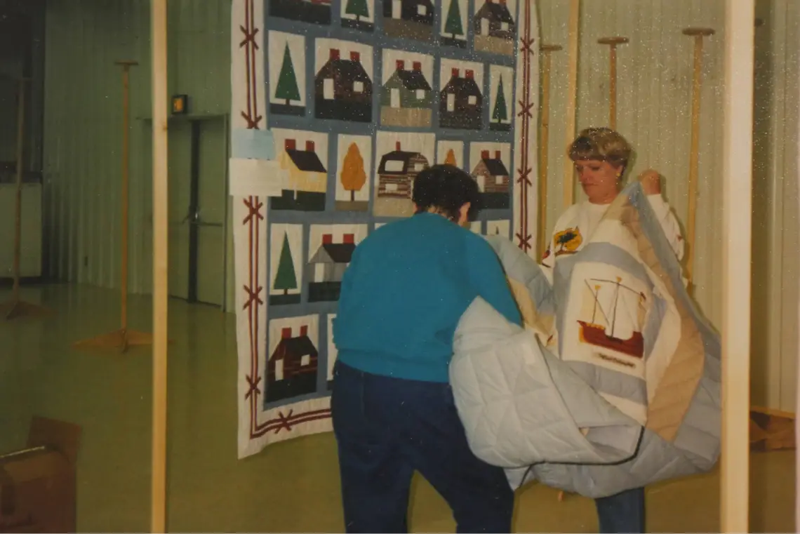 Two women hang a large quilt at an indoor quilting event, with another quilt displayed in the background.