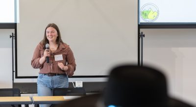 Peyton Loss stands in front of a whiteboard and screen with projected slide show, holding a microphone and facing the audience while giving a presentation