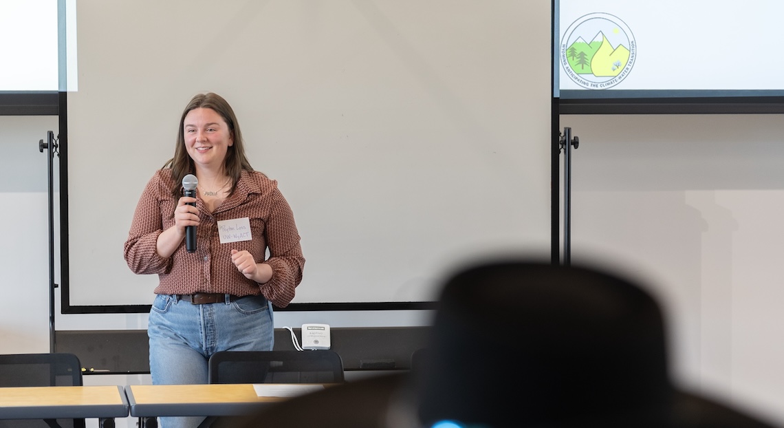 Peyton Loss stands in front of a whiteboard and screen with projected slide show, holding a microphone and facing the audience while giving a presentation