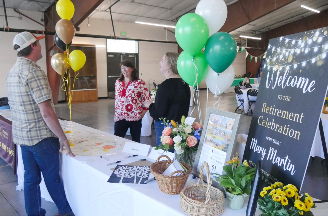 Mary Martin stands with guests near a decorated welcome table with balloons and a retirement celebration sign.