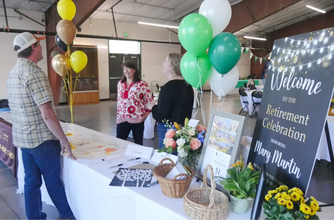 Mary Martin stands with guests near a decorated welcome table with balloons and a retirement celebration sign.