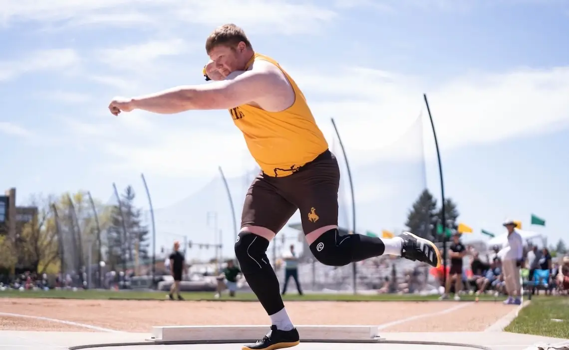 man wearing University of Wyoming track uniform competes in shotput at an outdoor track meet