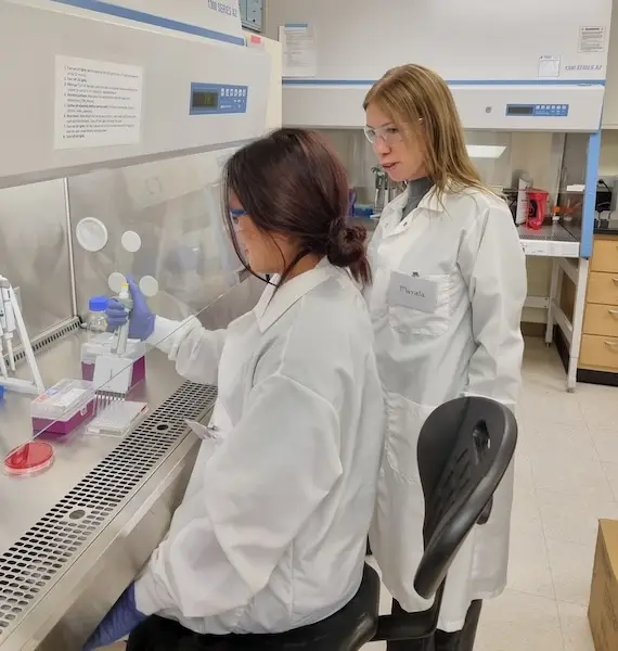 Jaiden Lopez and Mariela Srednik, dressed in white lab coats, stand beside a biosafety cabinet where Lopez, wearing safety gloves, performs a technical scientific procedure under a clear laboratory hood