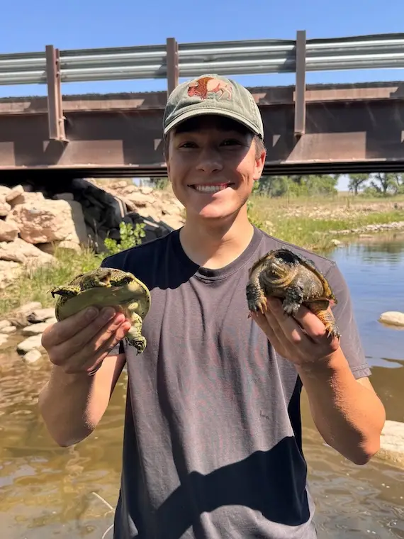 Camden Foley holds a turtle in each hand by a river.
